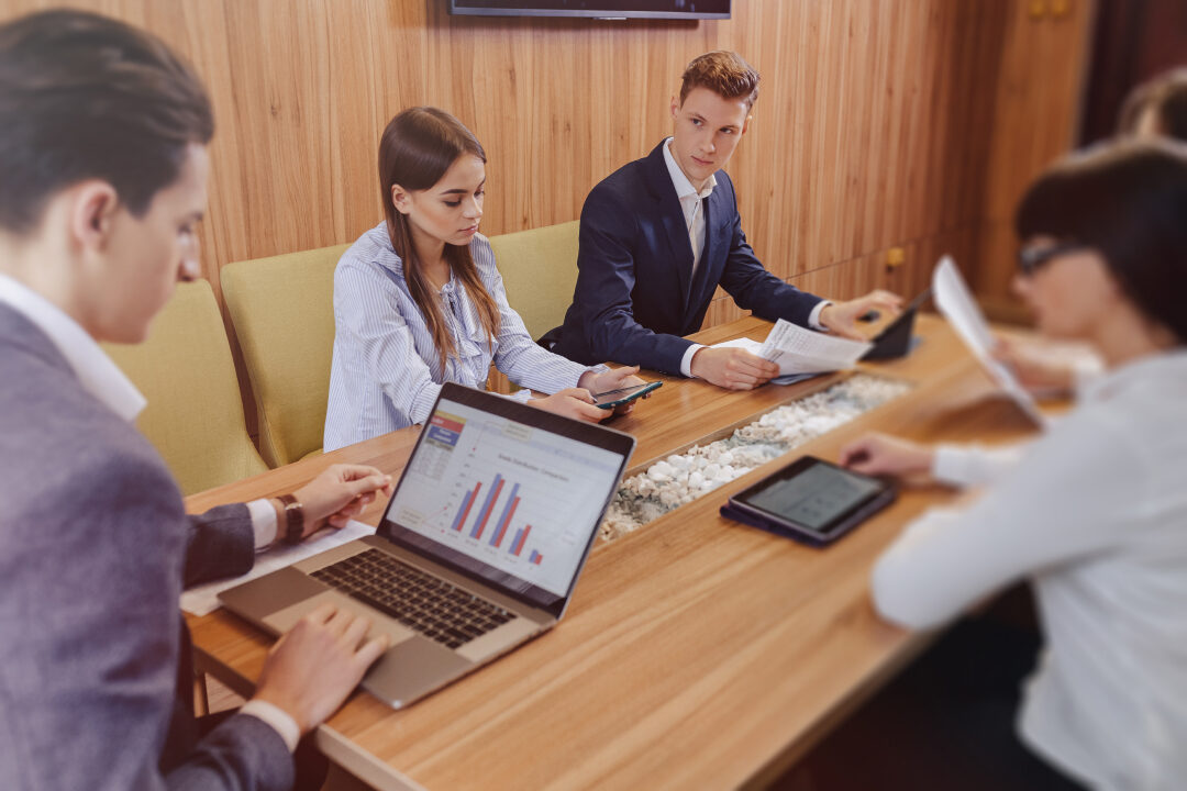 office-workers-hold-meeting-one-desk-laptops-tablets-papers-large-tv-set-wooden-wall